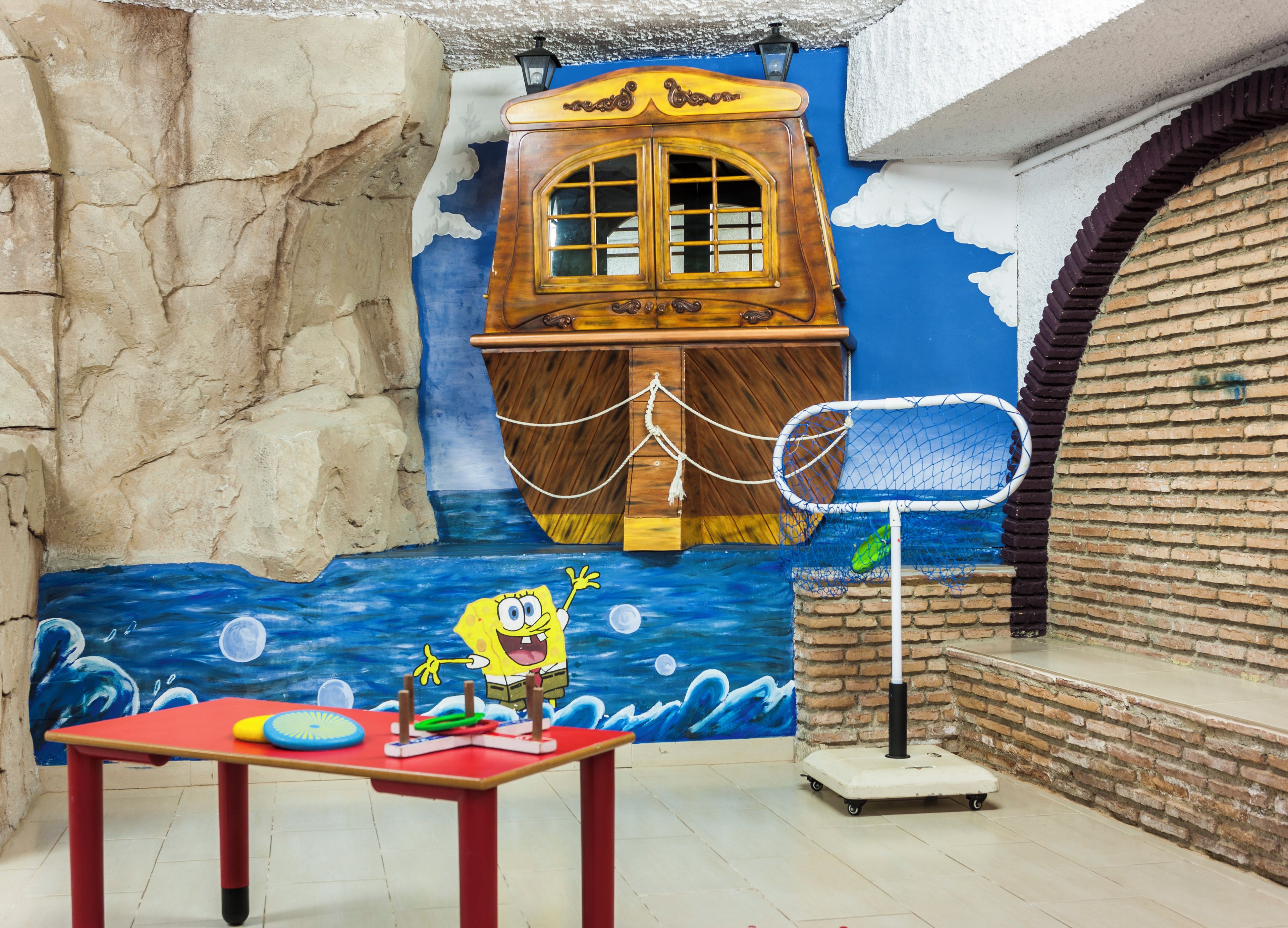 Habitación con un mural de un océano azul, un barco de madera y un personaje saludando. Se aprecia una mesa con juguetes y un aro de baloncesto.
