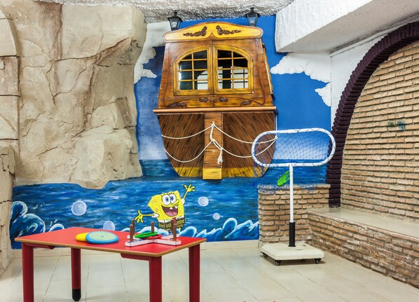 A room with a mural of a blue ocean, a wooden ship, and a character waving. There's a table with toys and a basketball hoop.