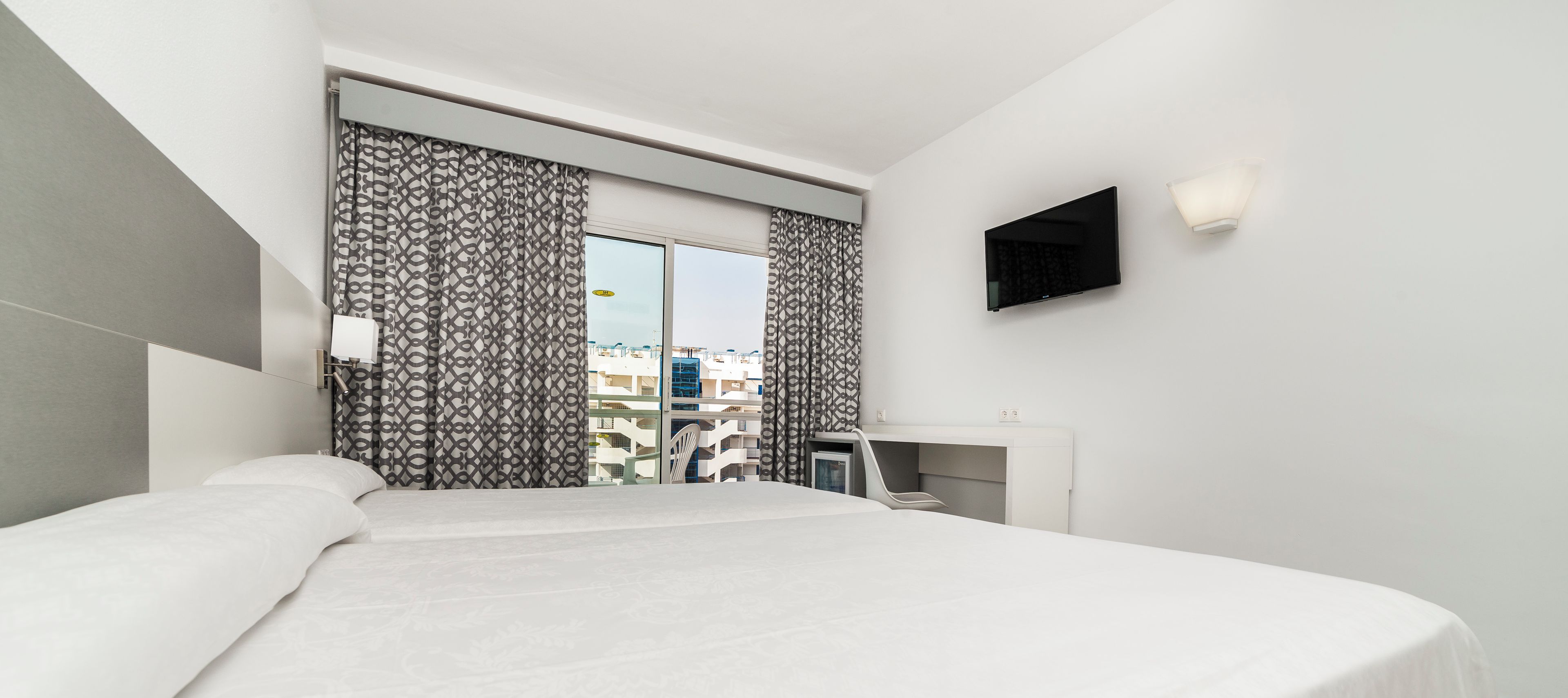 A modern hotel room with a large bed, a desk, a flat-screen TV, and a view of the city through the window.