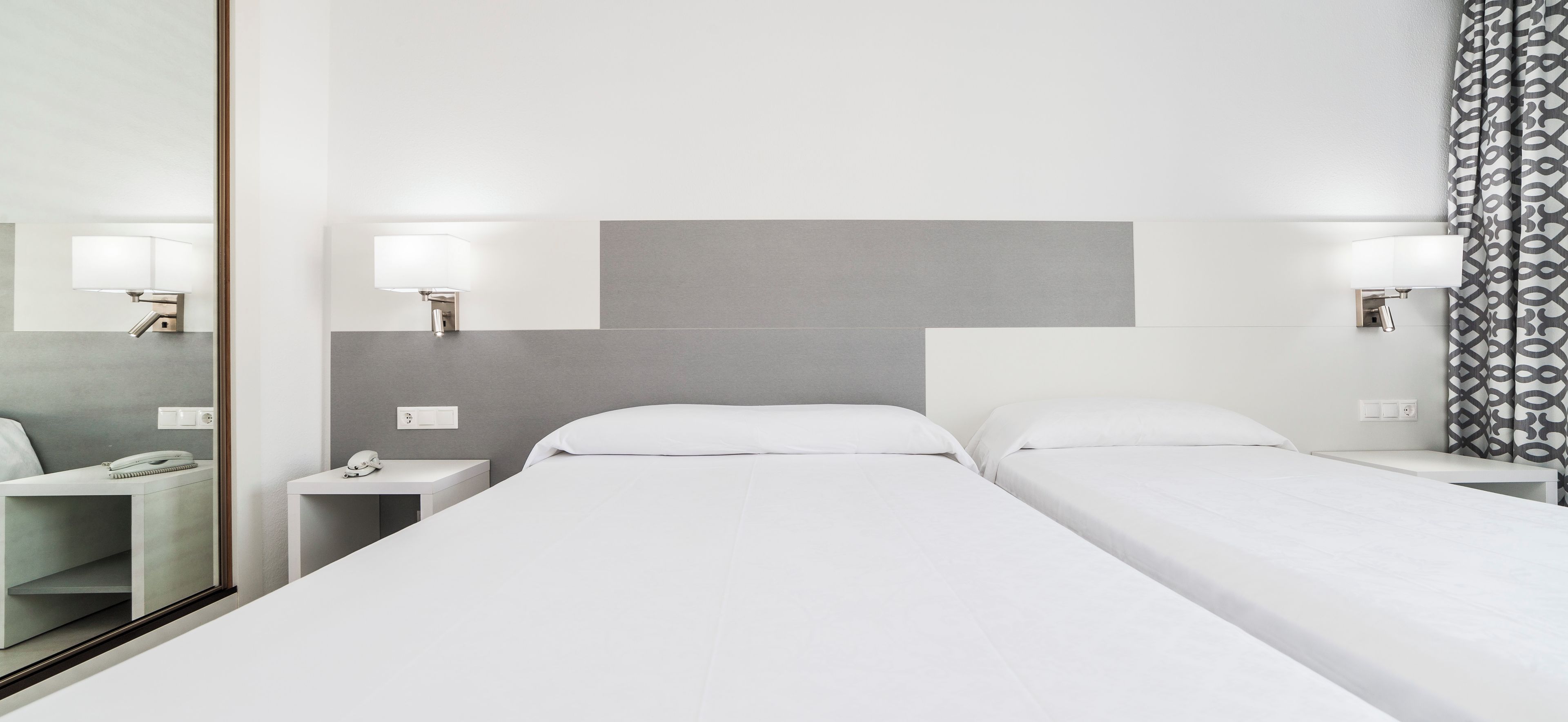 Habitación de hotel con dos camas individuales, una mesita de noche con lámpara, un espejo y una cortina estampada.