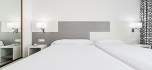 Habitación de hotel con dos camas individuales, una mesita de noche con lámpara, un espejo y una cortina estampada.