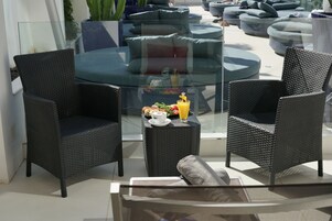 A modern outdoor seating area with wicker chairs and a glass table, featuring a breakfast setup with a beverage and pastries.