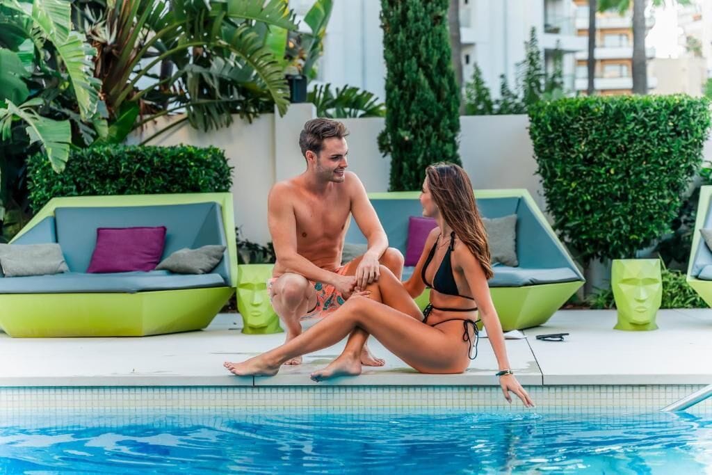 Un couple est assis au bord d’une piscine, avec des meubles modernes et de la verdure en arrière-plan.