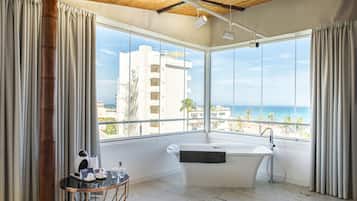 A modern bathroom with a freestanding bathtub, large windows offering a view of the beach and buildings, and a small table with toiletries.
