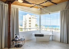 Un baño moderno con una bañera independiente, ventanas amplias con vistas a la playa y edificios, y una mesita con artículos de aseo.