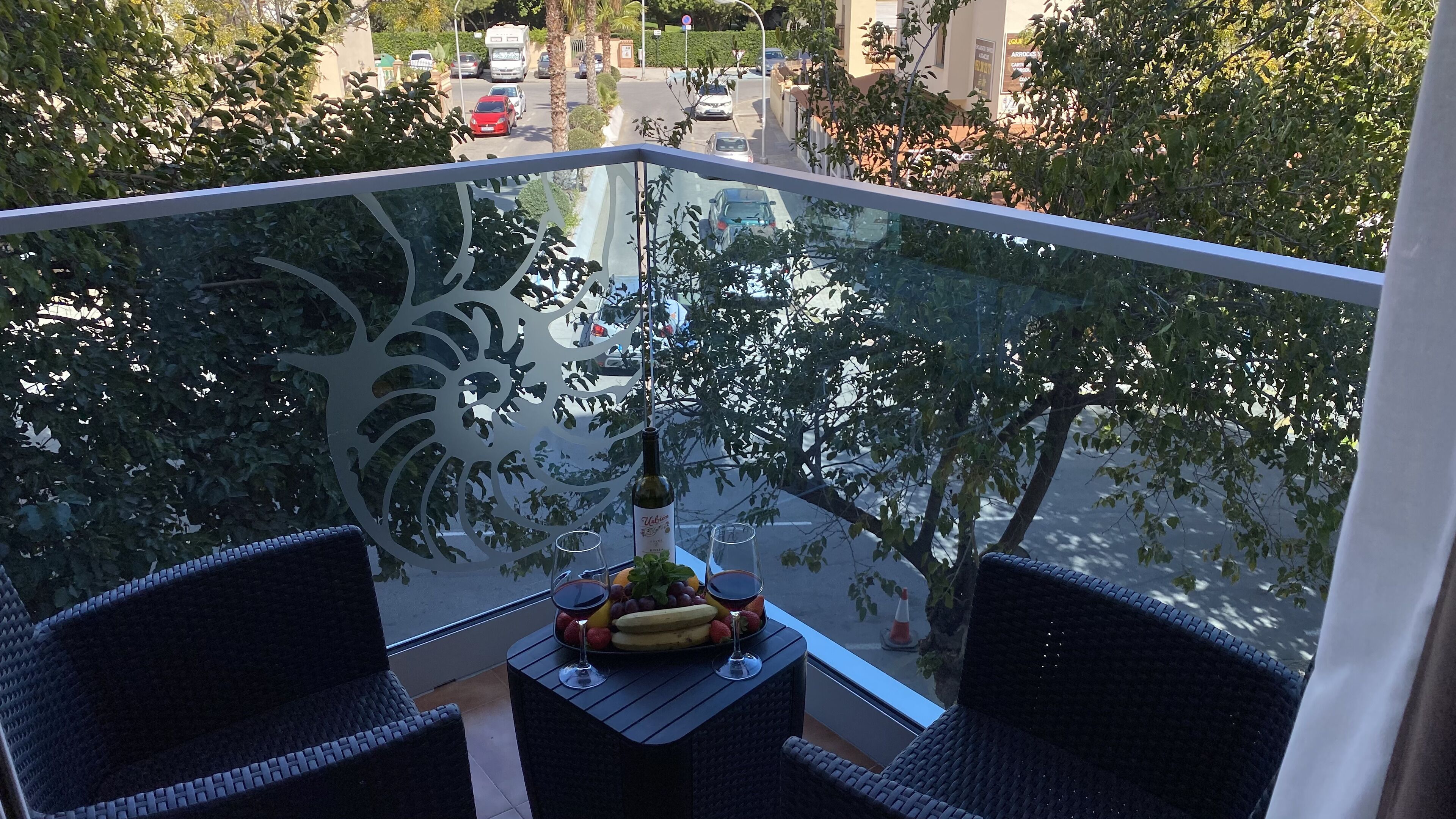 A balcony with a glass railing, two wicker chairs, a small table with a bottle of wine, a fruit platter, and a view of a street with cars and trees.