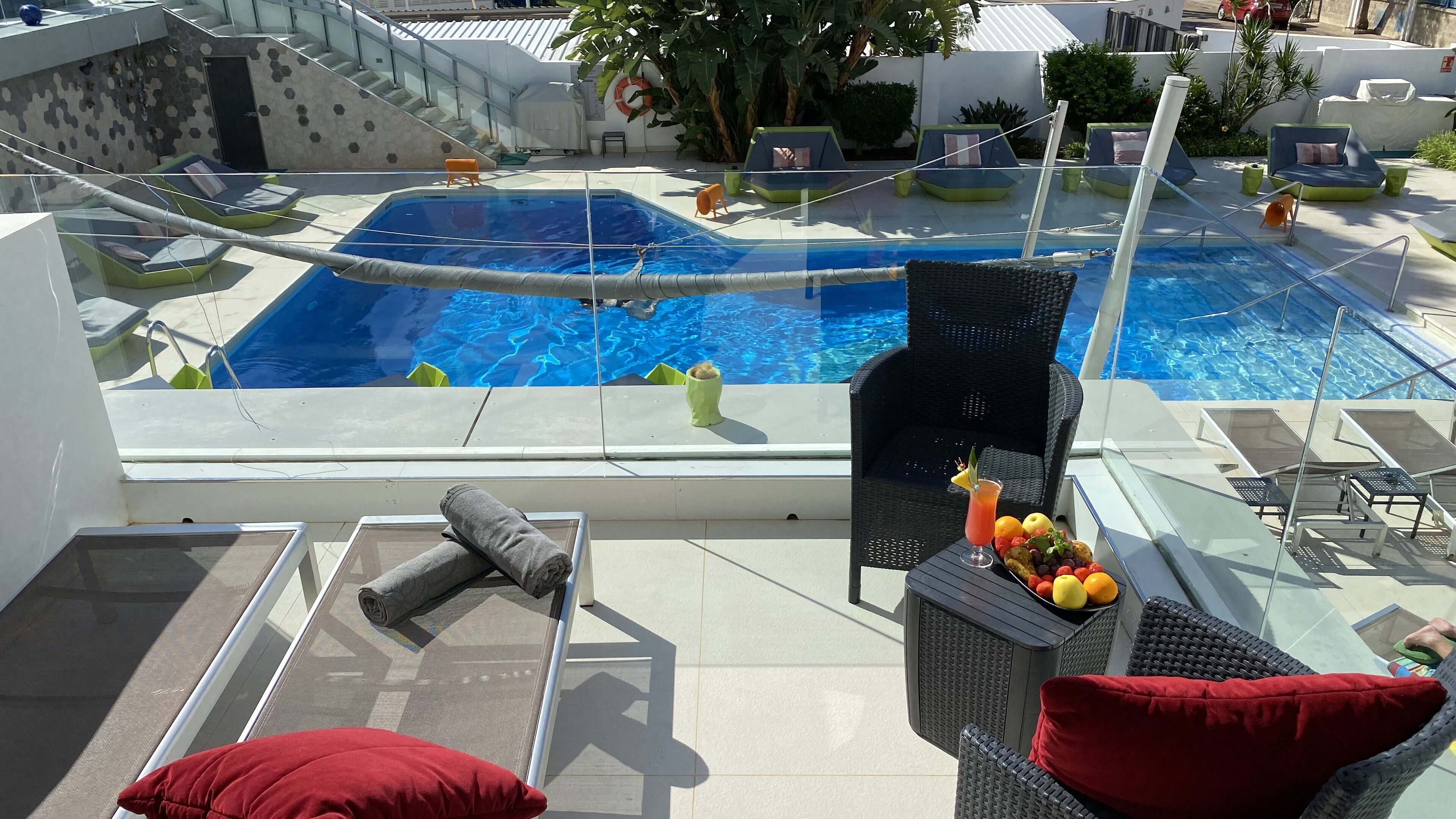 A rooftop pool area with lounge chairs, a small table with fruit and a drink, and a view of the pool.