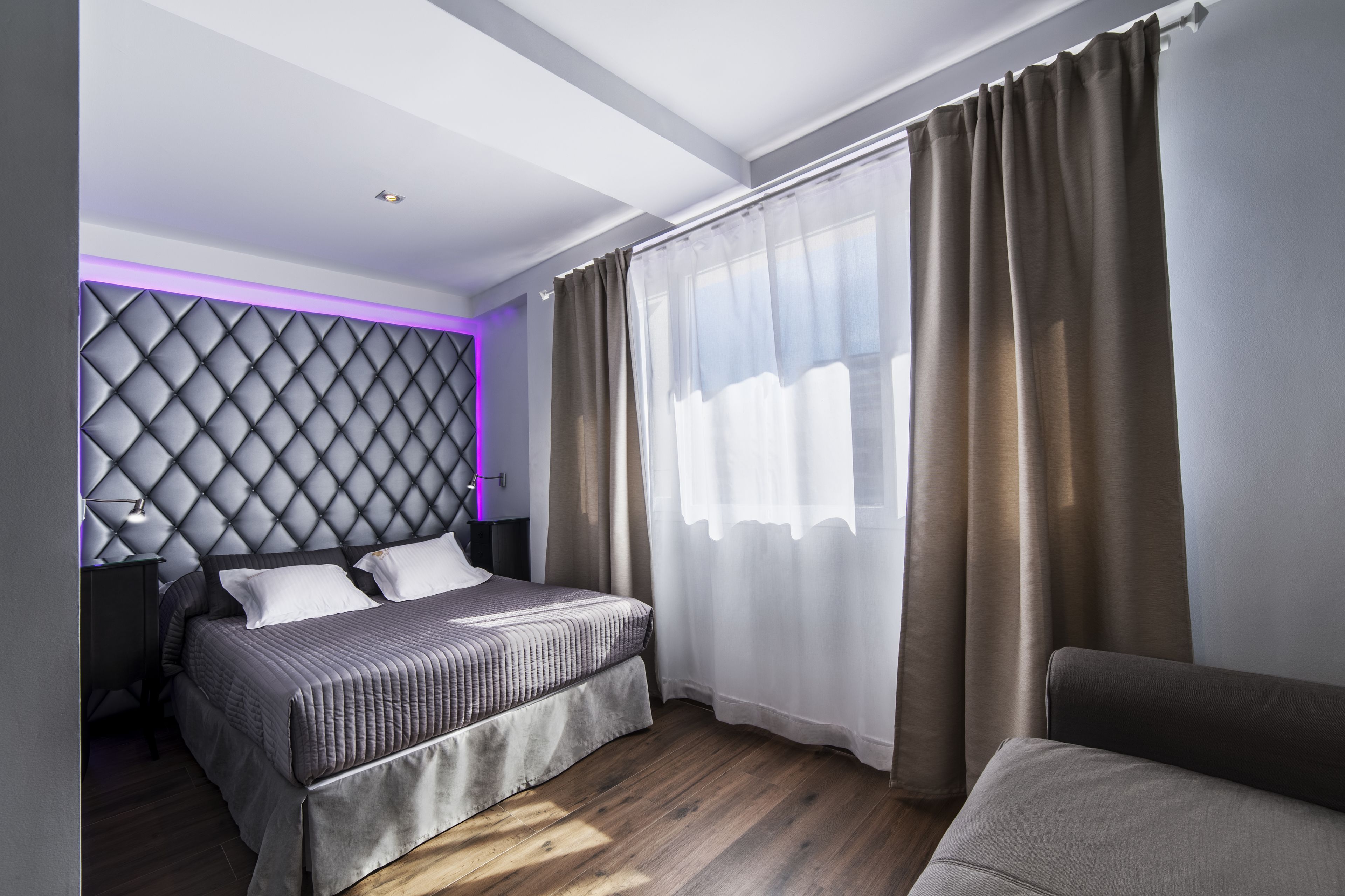 Un dormitorio moderno con un cabecero capitonado, una cama con colcha gris, una mesita de noche y un ventanal con cortinas de gasa.