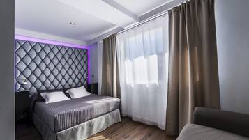 Un dormitorio moderno con un cabecero capitonado, una cama con colcha gris, una mesita de noche y un ventanal con cortinas de gasa.