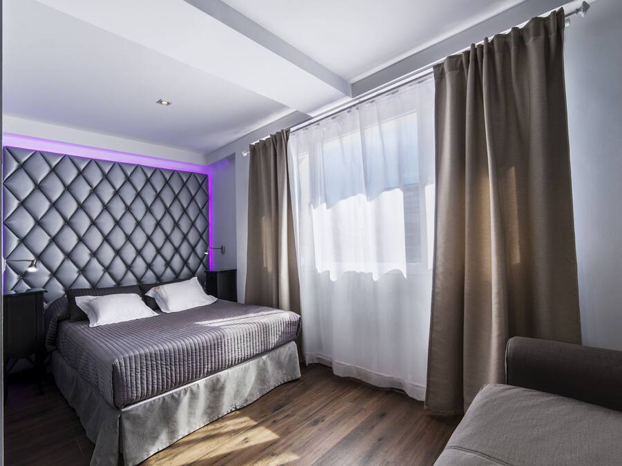 A modern bedroom with a tufted headboard, a bed with a grey quilt, a bedside table, and a large window with sheer curtains.