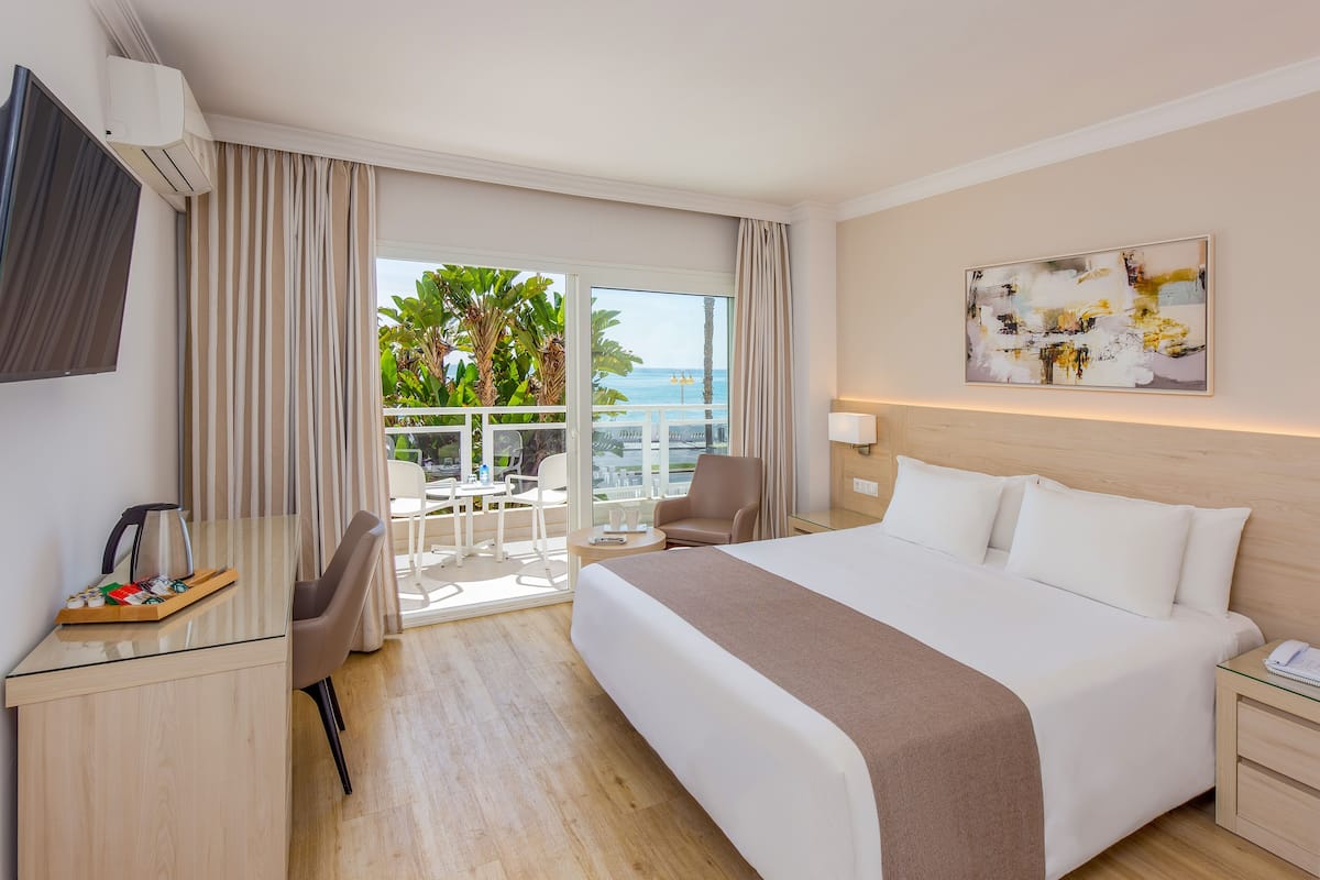 A hotel room with a large bed, a desk with a coffee maker, a chair, a balcony with a view of the sea, and a painting on the wall.