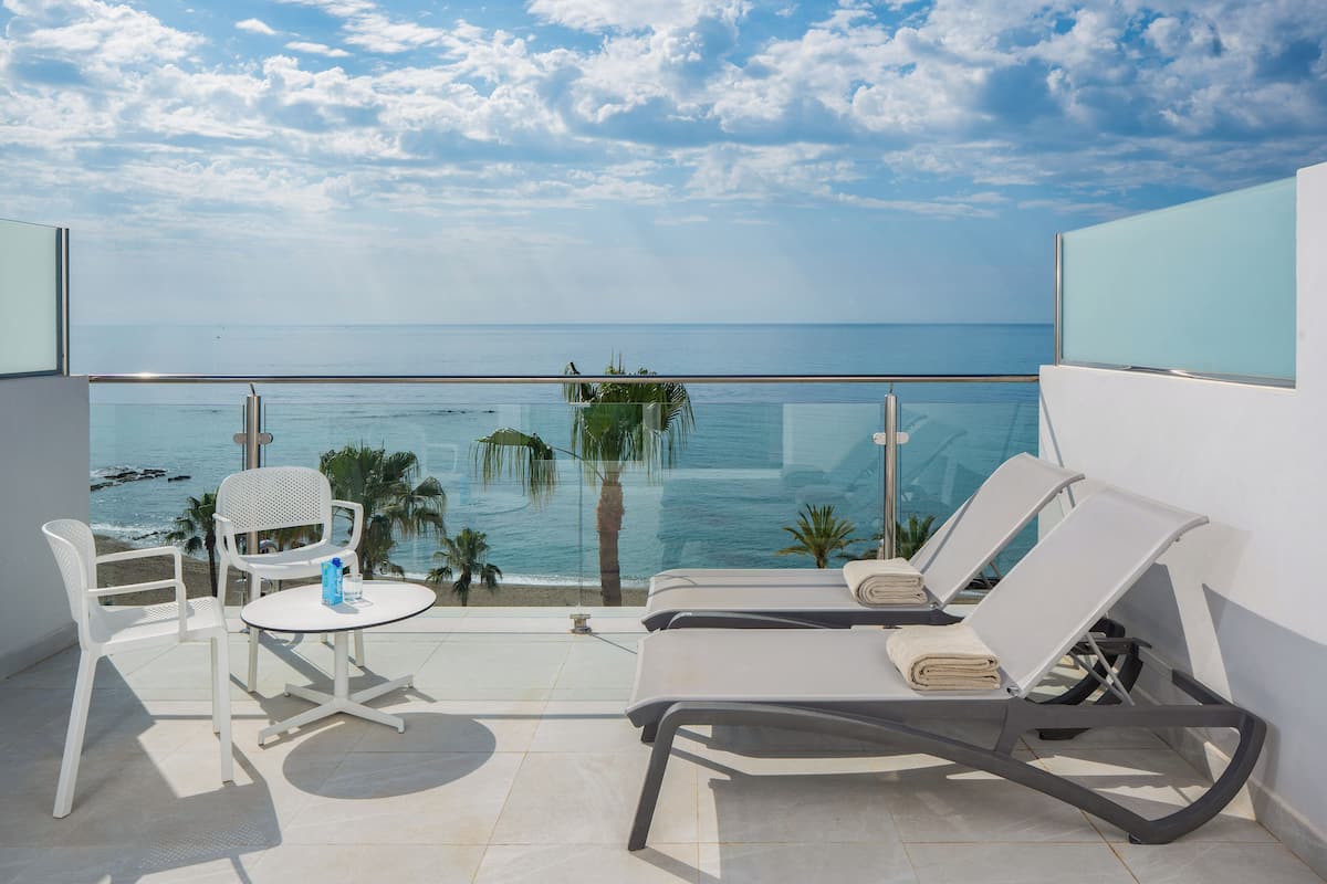 A balcony with a glass railing, white lounge chairs, a small table, and a view of the ocean.