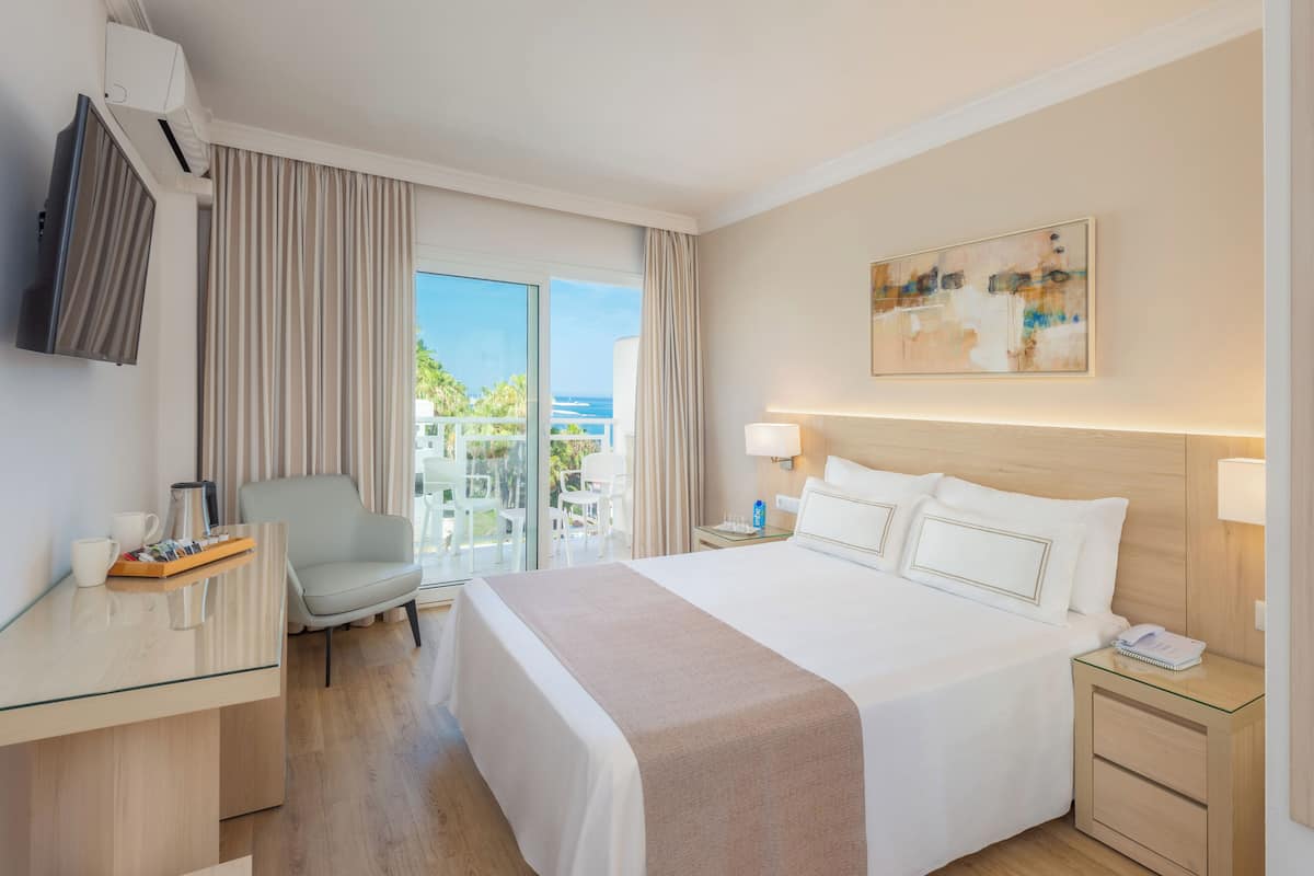 A hotel room with a large bed, a desk with a chair, a TV, a balcony with a view of the ocean, and a painting on the wall.