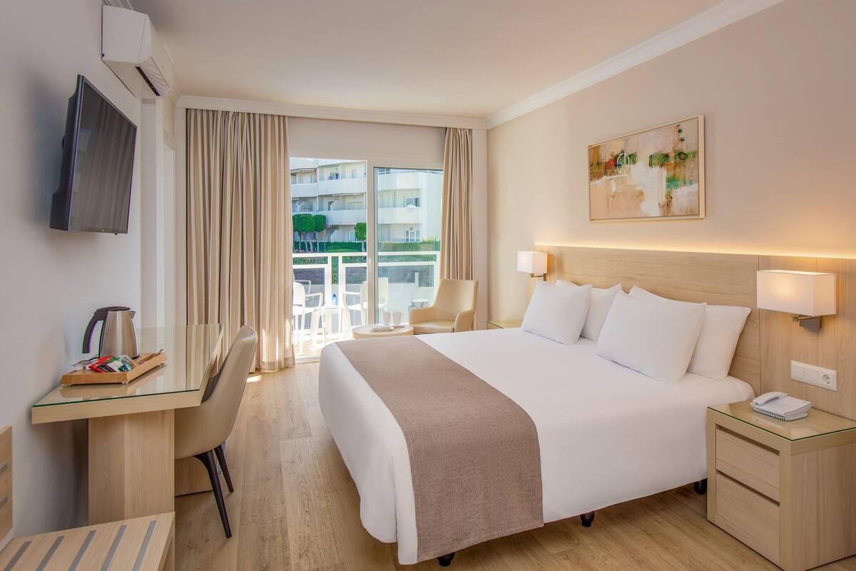 A hotel room with a bed, bedside tables, a desk with a chair, a TV, and a large window with curtains.