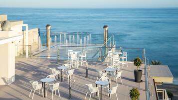 Terraço no telhado com cadeiras e mesas brancas, vista para o mar.