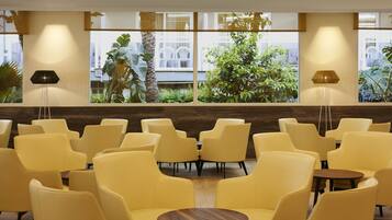 A modern indoor seating area with yellow chairs and tables, large windows with a view of greenery and buildings, and two floor lamps.