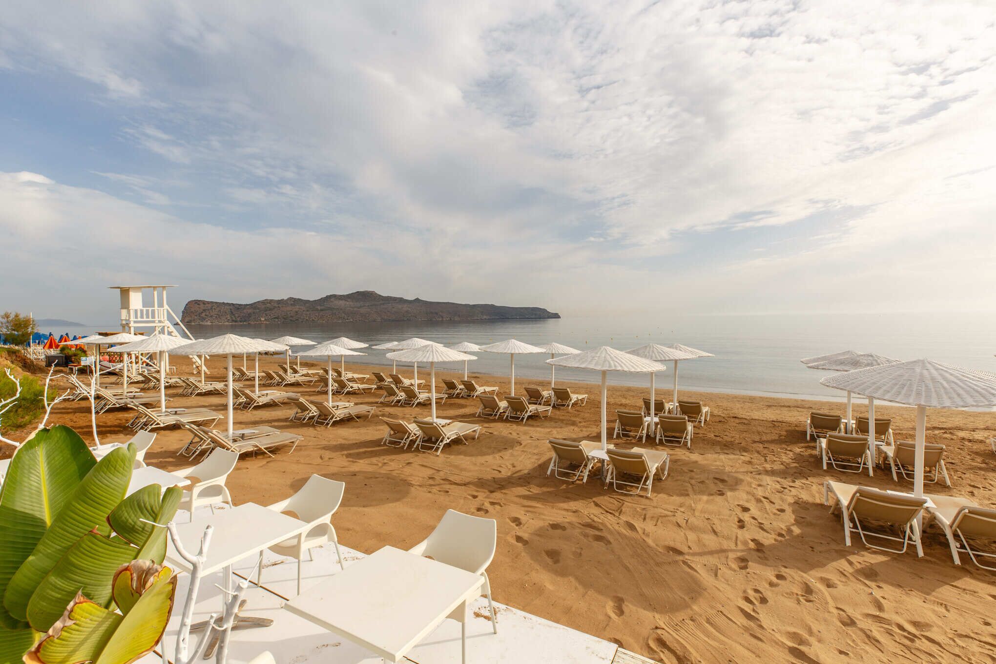 Una spiaggia con sedie a sdraio e ombrelloni bianchi, una torretta di salvataggio e vista sul mare e sulle colline lontane.