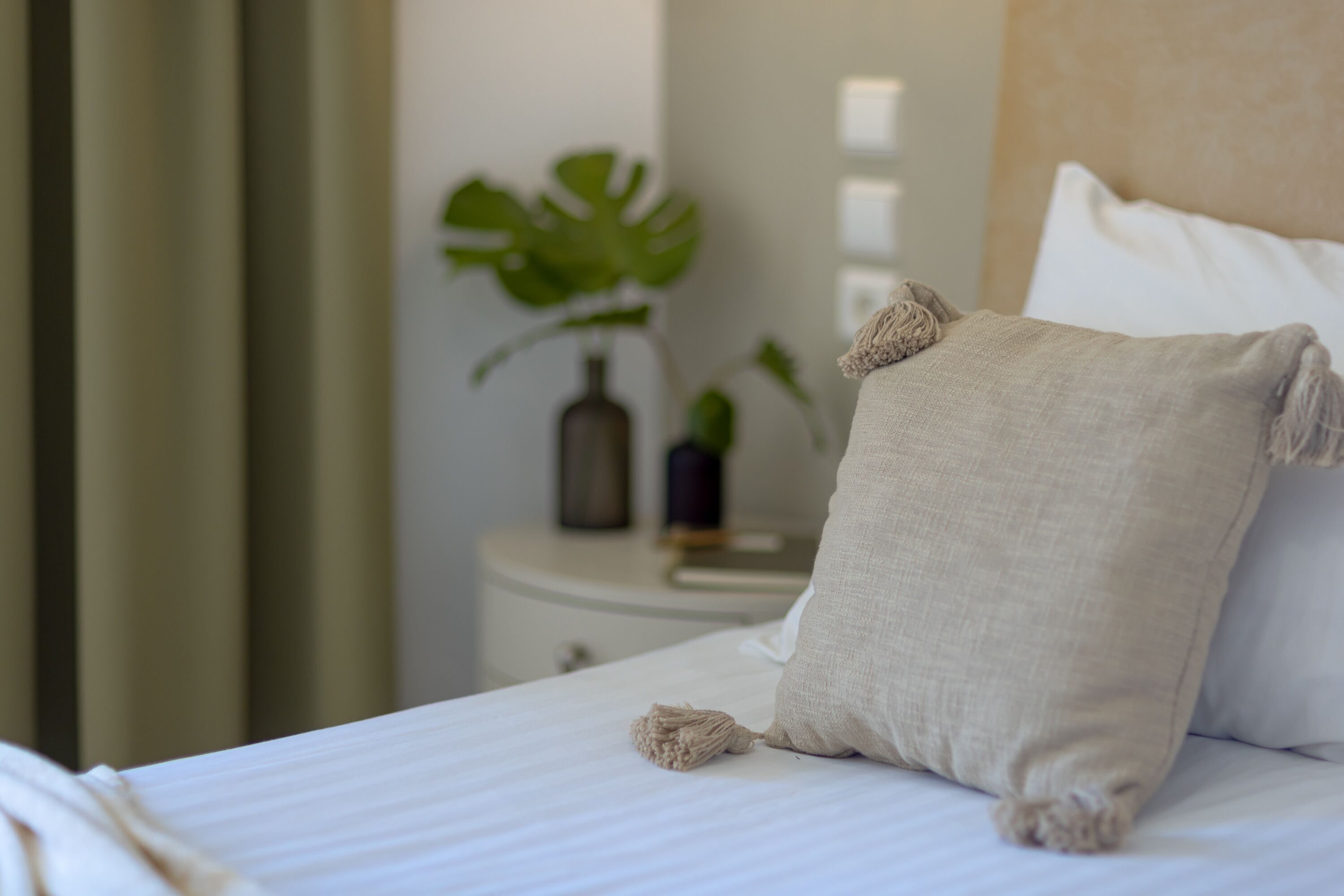 Cama bem arrumada com travesseiro branco e almofada bege. Ao fundo, uma mesinha redonda com uma planta em vaso.