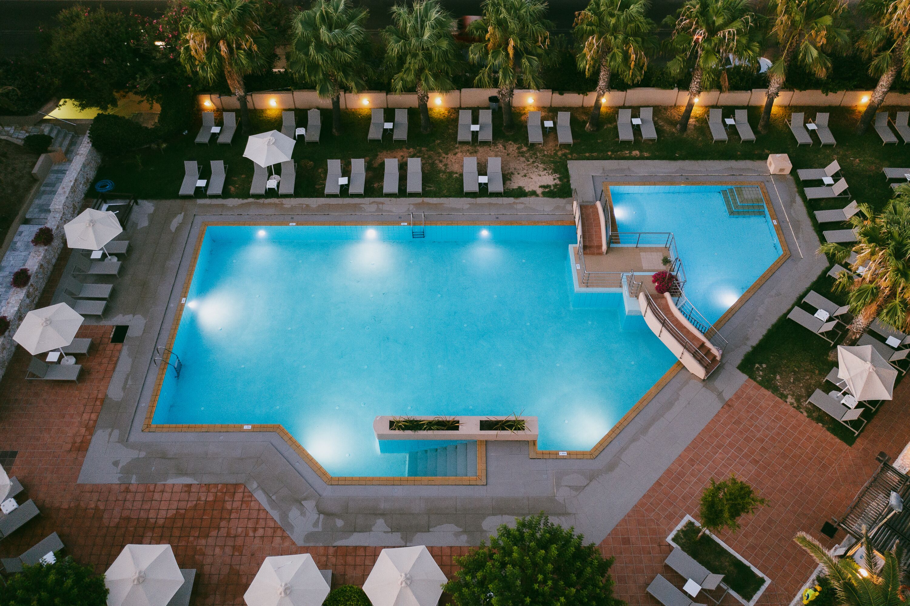 An aerial view of a rectangular swimming pool with a diving board, surrounded by lounge chairs and umbrellas.