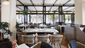 Een restaurant met hoge plafonds, houten balken en grote ramen. Tafels zijn gedekt met witte tafelkleden en stoelen.