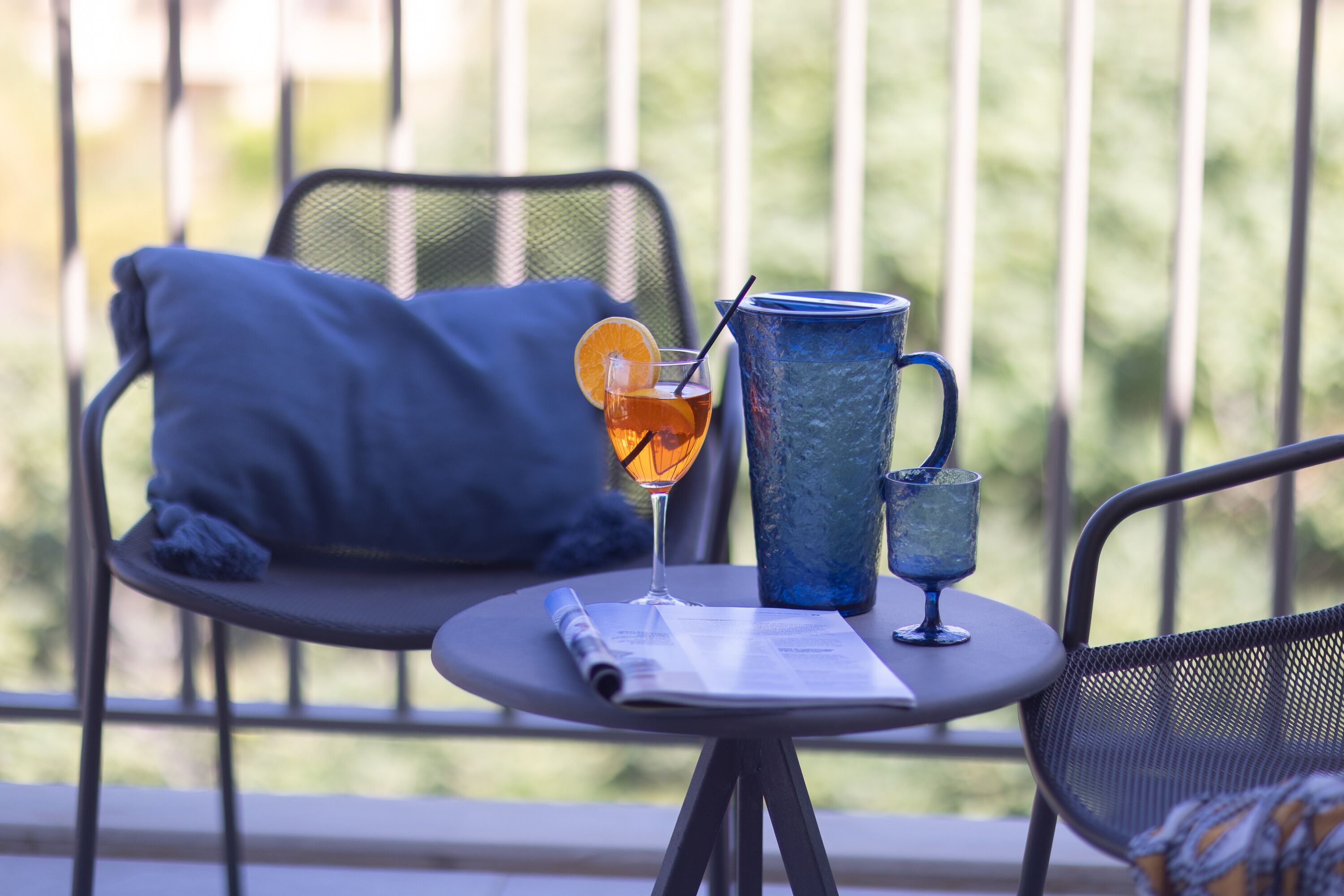 Un balcon avec une table, deux chaises, un coussin bleu, un verre avec une paille et un menu.