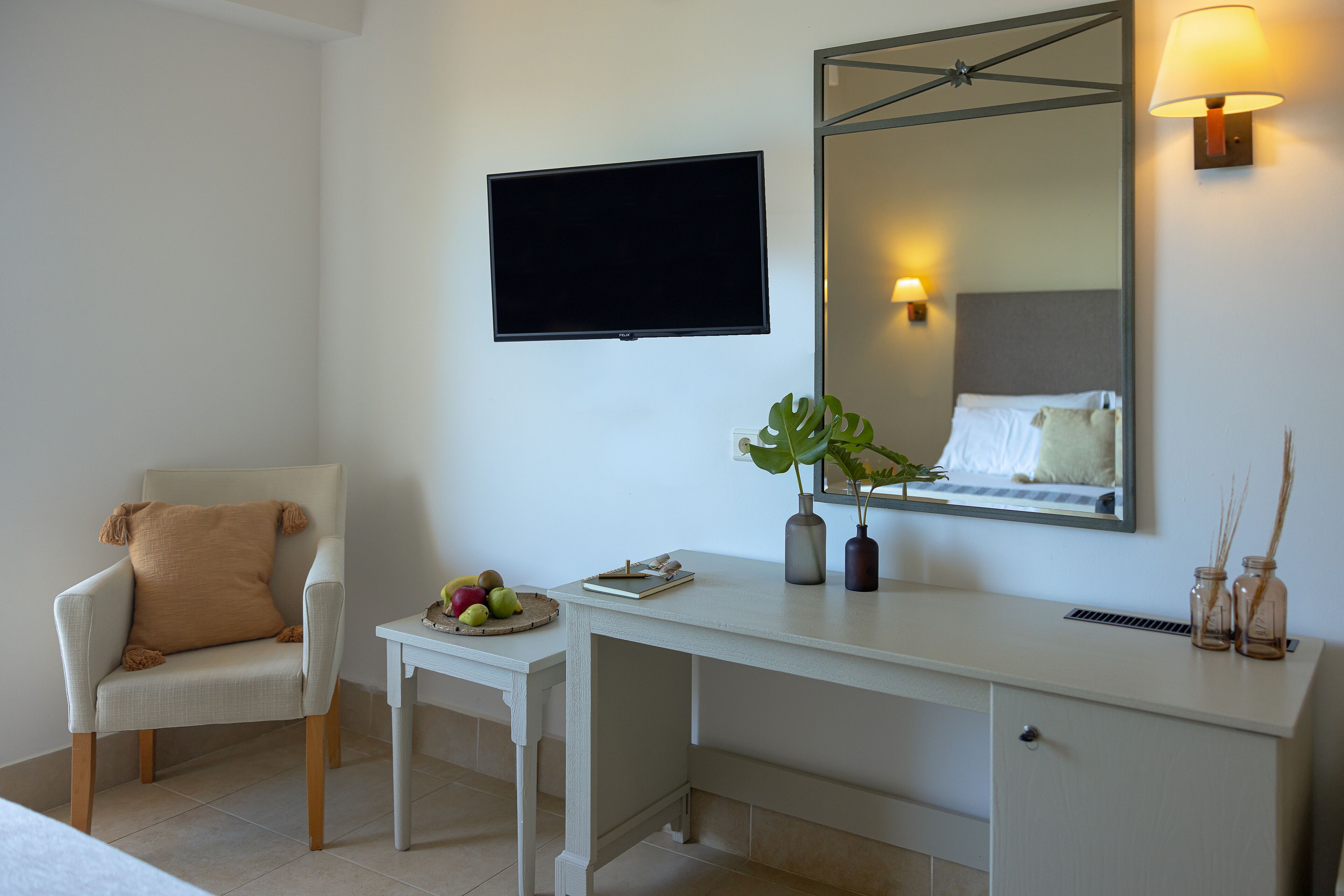 A hotel room with a white desk, a chair, a flat-screen TV, a mirror, and a bedside table with a plant and a fruit bowl.