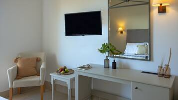 A hotel room with a white desk, a chair, a flat-screen TV, a mirror, and a bedside table with a plant and a fruit bowl.