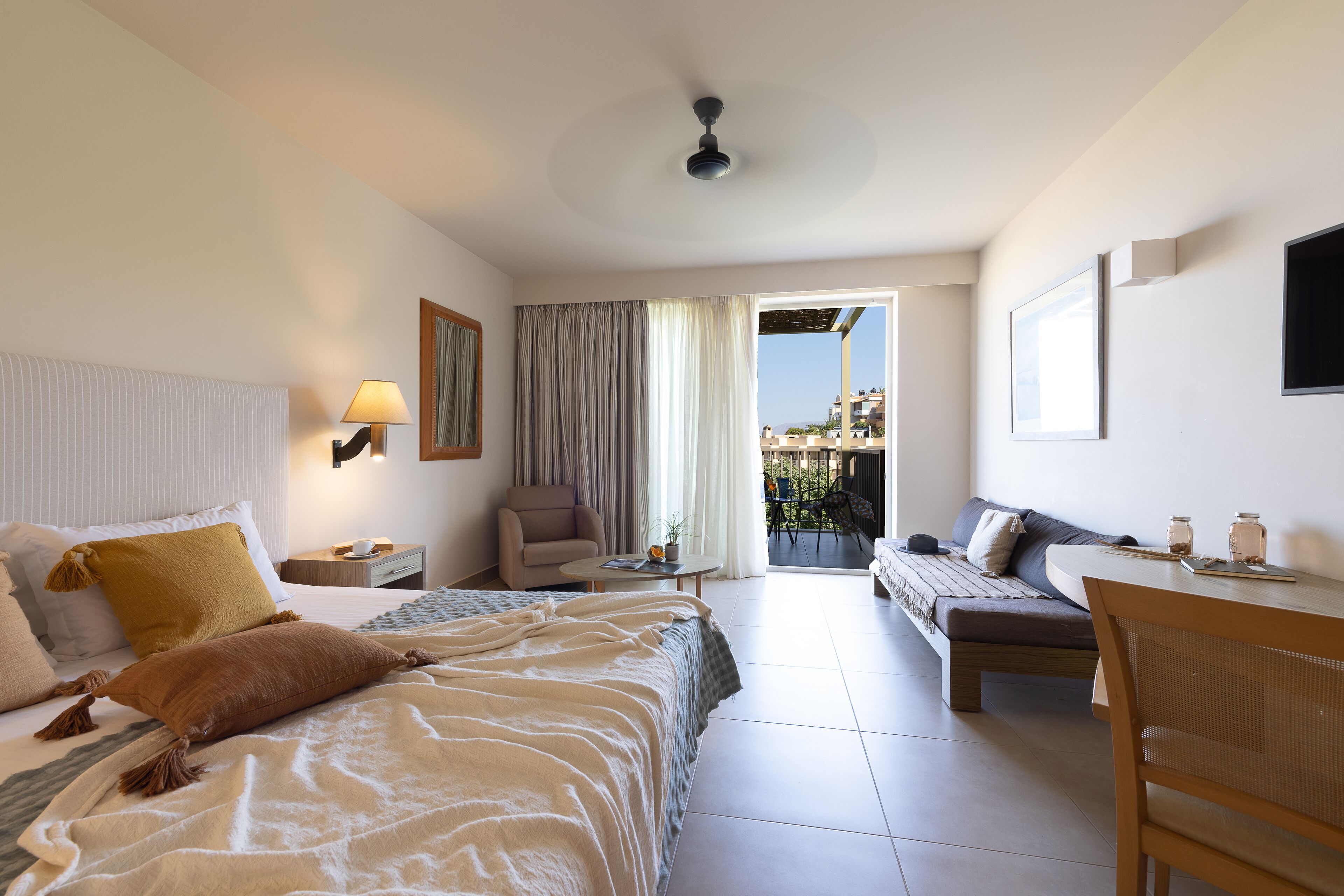 Ein Hotelzimmer mit Bett, Sofa, Esstisch und Stuhl. Es gibt ein Fenster mit Vorhängen, einen Deckenventilator und Blick auf Gebäude.