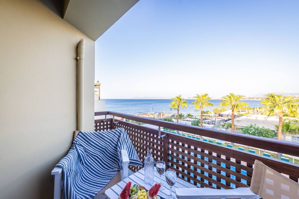 Un balcón con vista al mar, una mesa con una botella, vasos y fruta, y un sofá a rayas.