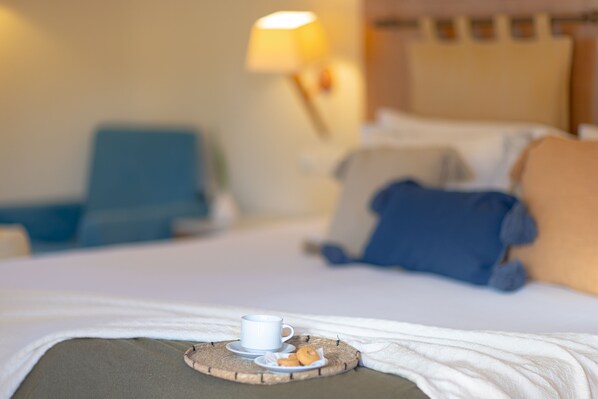 A neatly made bed with a cup and pastries on a tray, a lamp, and a chair in a hotel room. - Giannoulis Santa Marina Plaza - Adults Only (Chania)