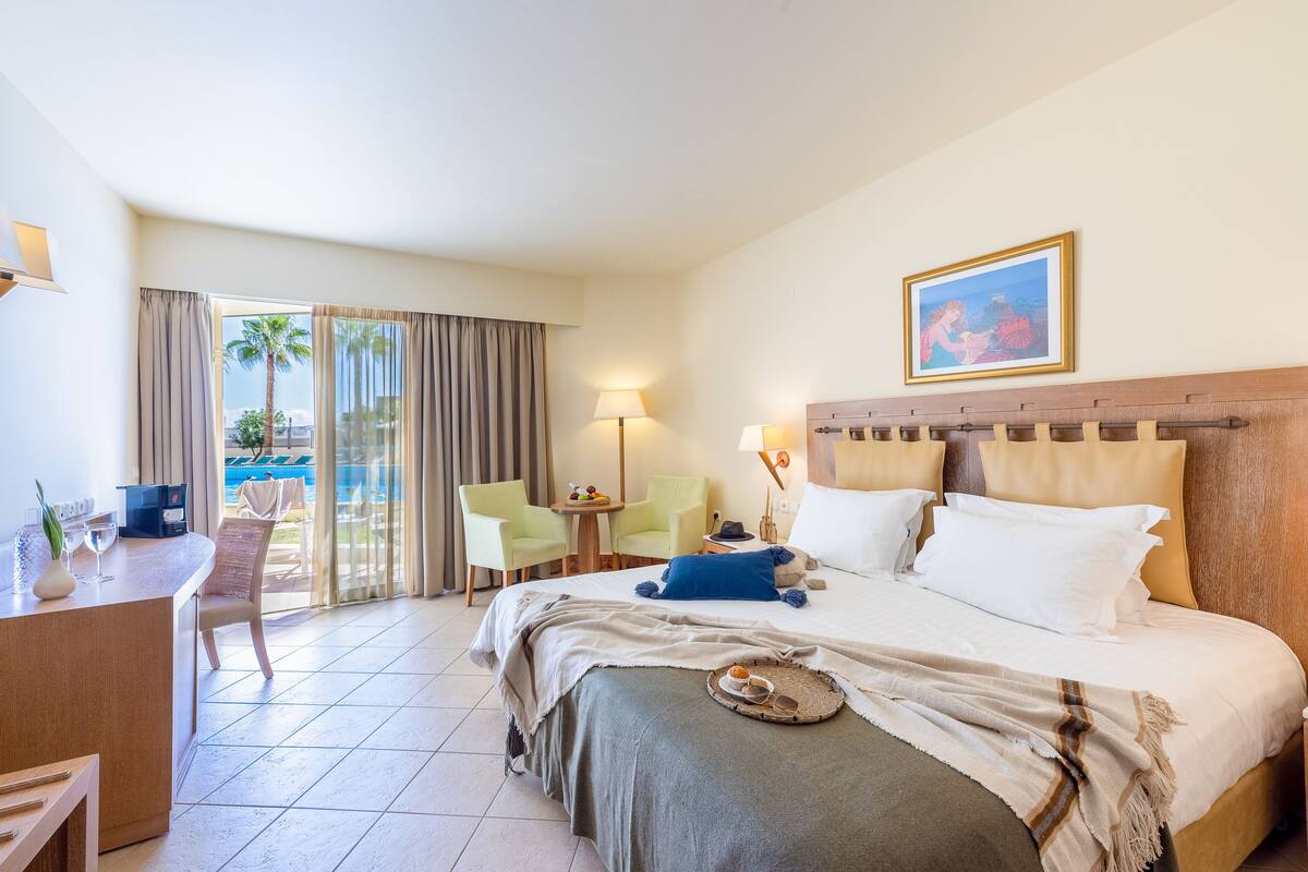 Habitación de hotel con cama, escritorio, sillas, una mesita y vista a la piscina.