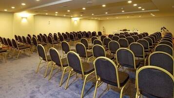 Sala de conferências com fileiras de cadeiras e mesas, piso coberto de carpete e iluminação embutida.