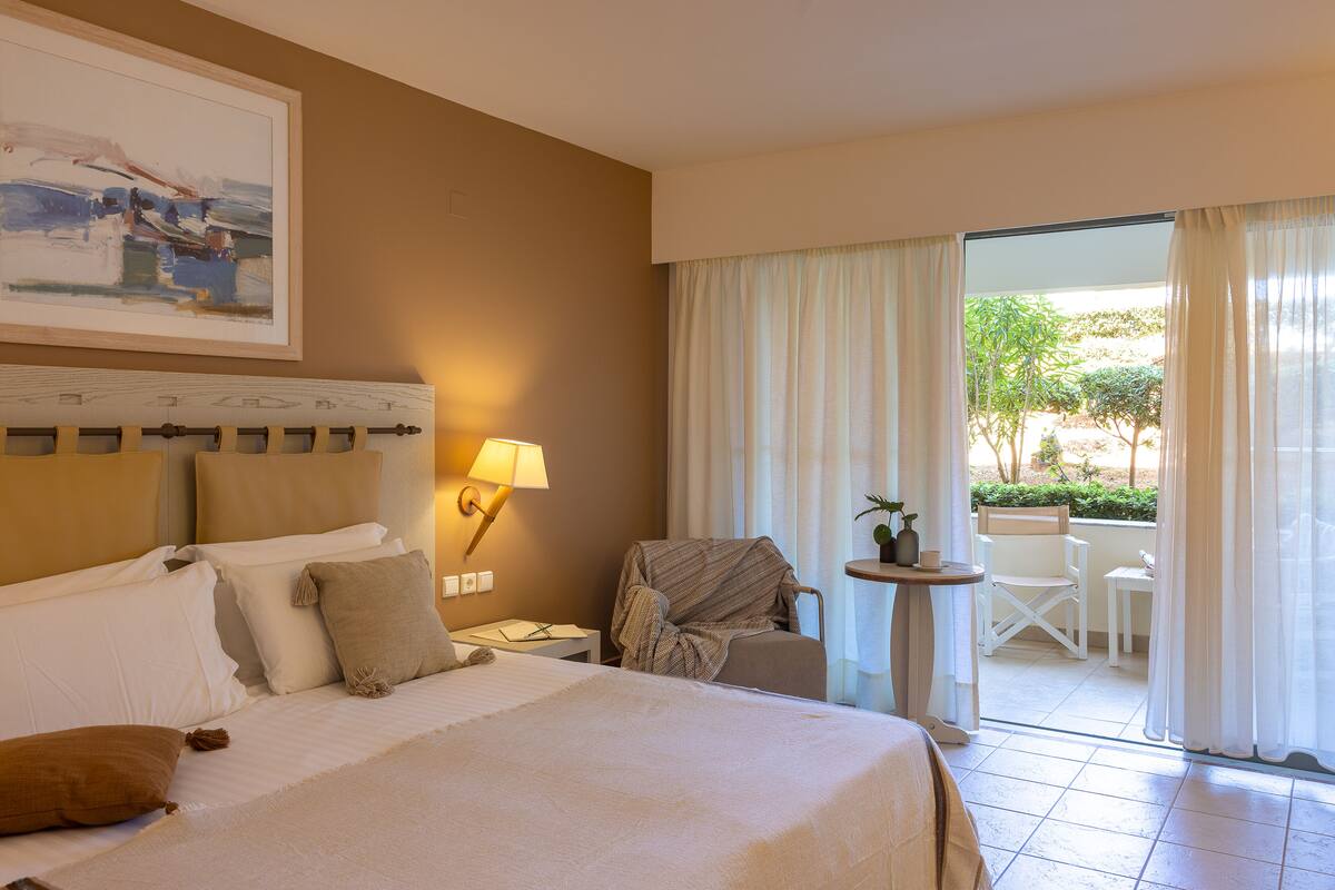 Habitación de hotel con cama, mesita de noche, lámpara, silla y vistas al exterior.
