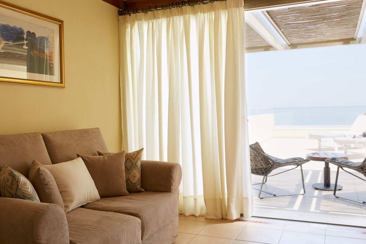 Un sofá color beige con cojines estampados, una cortina blanca y vista al mar desde un balcón con mobiliario exterior.