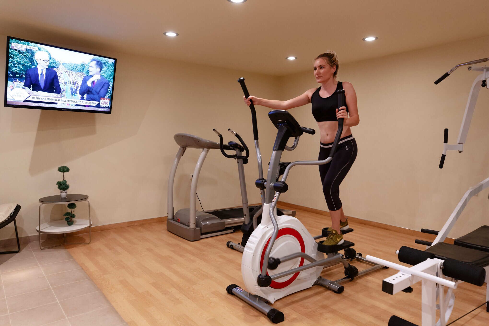A woman exercising on an elliptical trainer in a room with a television displaying a news program, a small table with plants, and a bench.