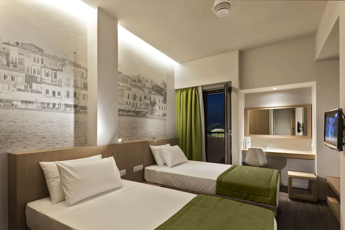 Habitación de hotel con dos camas, un escritorio, una silla, un televisor y un ventanal grande con cortinas.