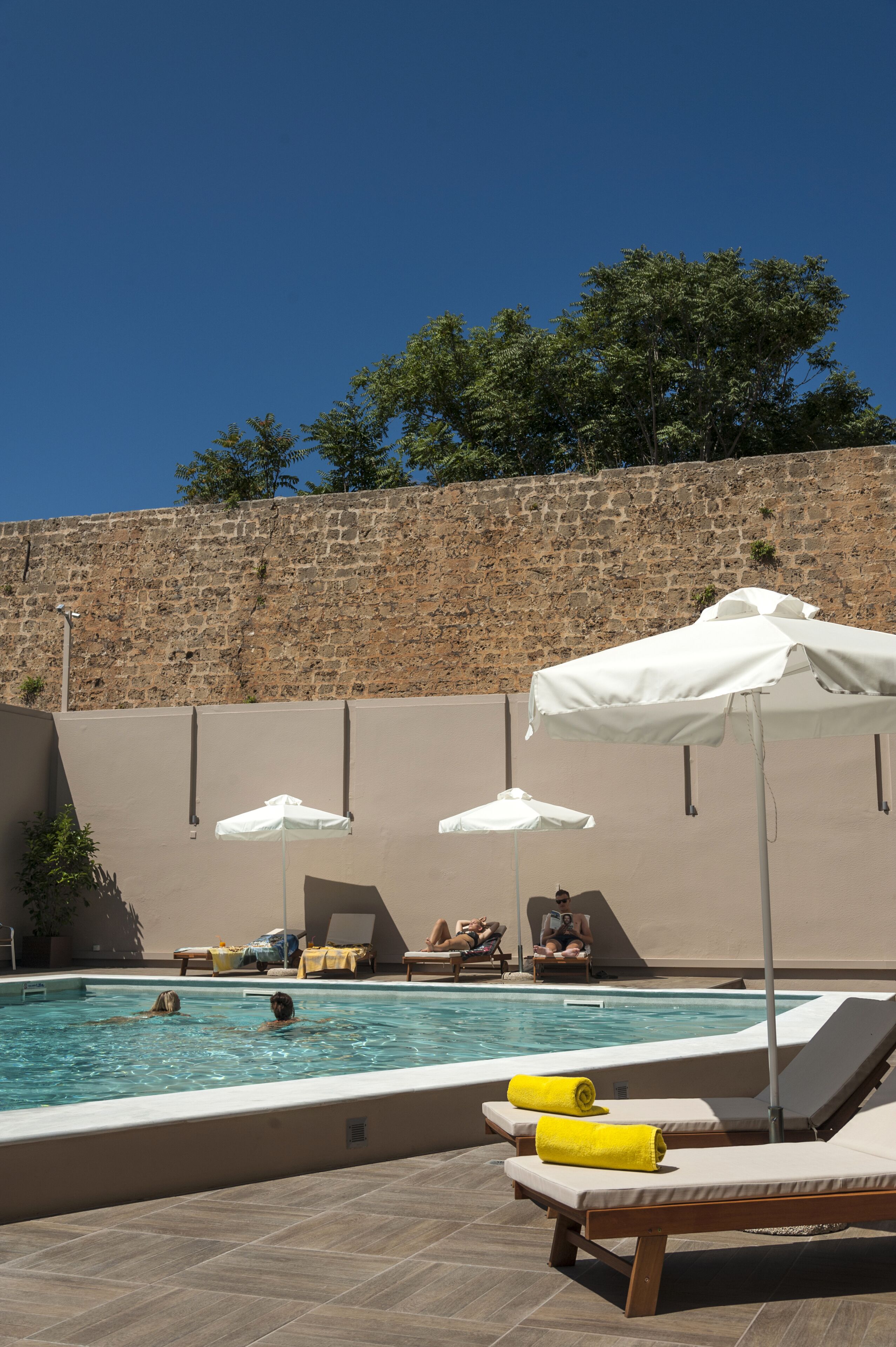 A swimming pool with lounge chairs and umbrellas, a stone wall, and trees in the background.