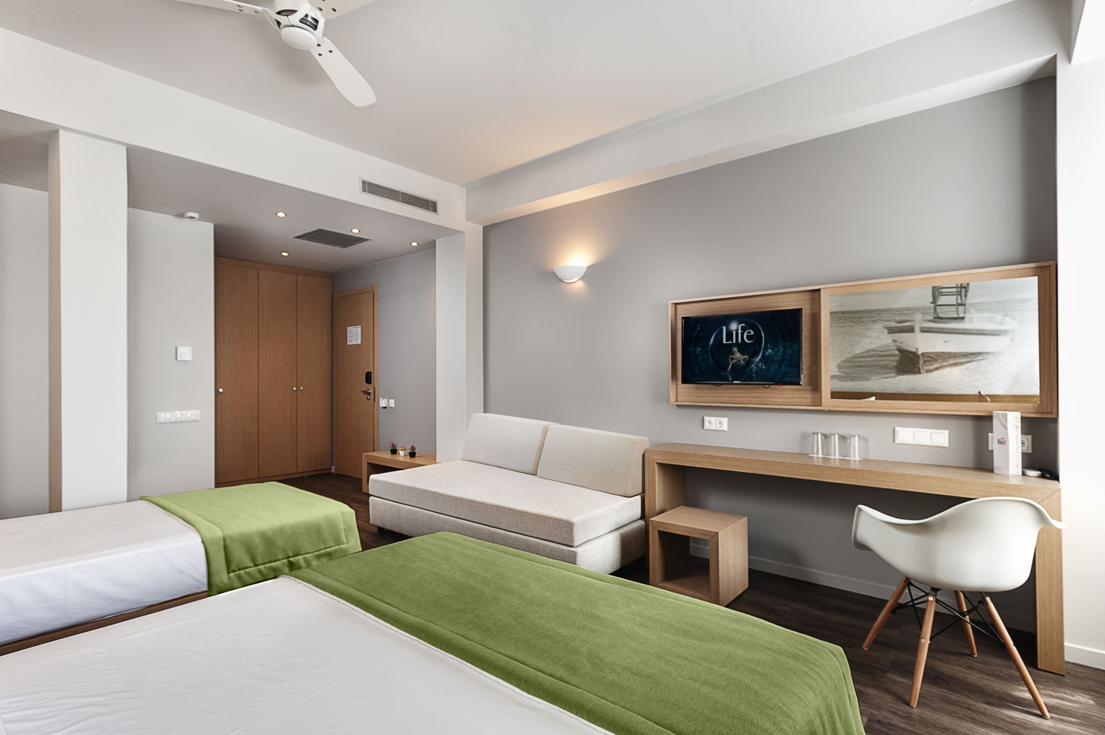 Una habitación de hotel moderna con una cama sofa, un escritorio con una silla, un ventilador de techo y obras de arte en la pared.
