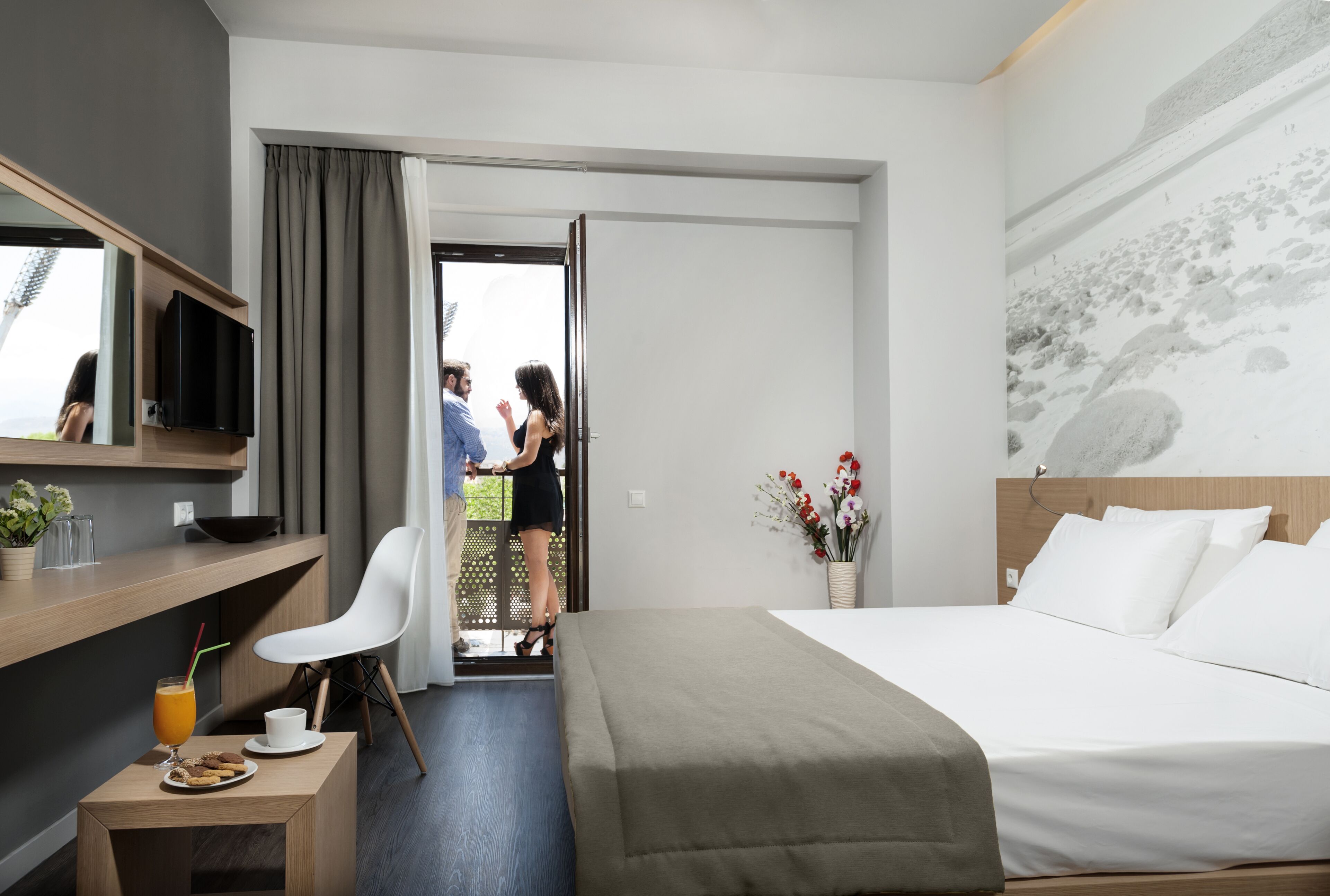 Quarto de hotel com cama, escrivaninha com xícara e biscoitos, cadeira, televisão e vista para o exterior.
