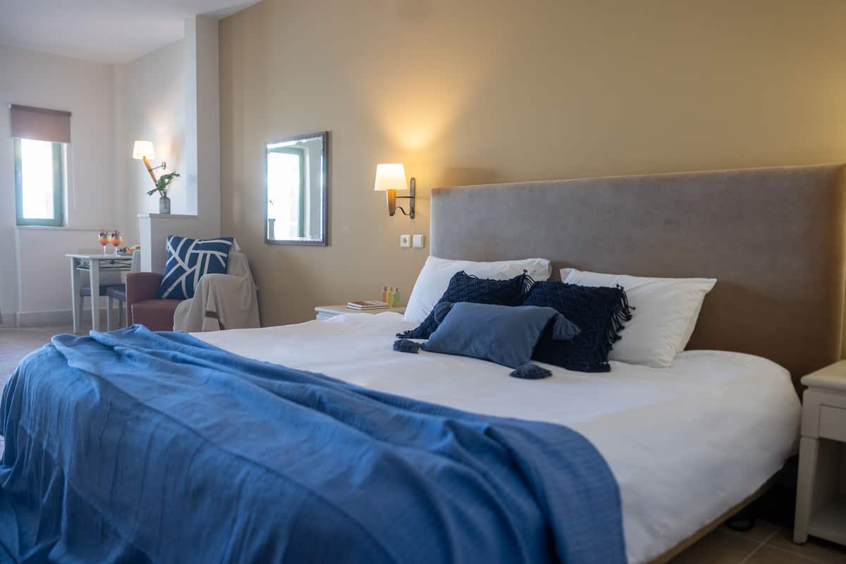 Ein Hotelzimmer mit einem großen Bett, einem Nachttisch, einem Sessel, einem Schreibtisch mit Lampe und einem fenster mit Vorhängen.