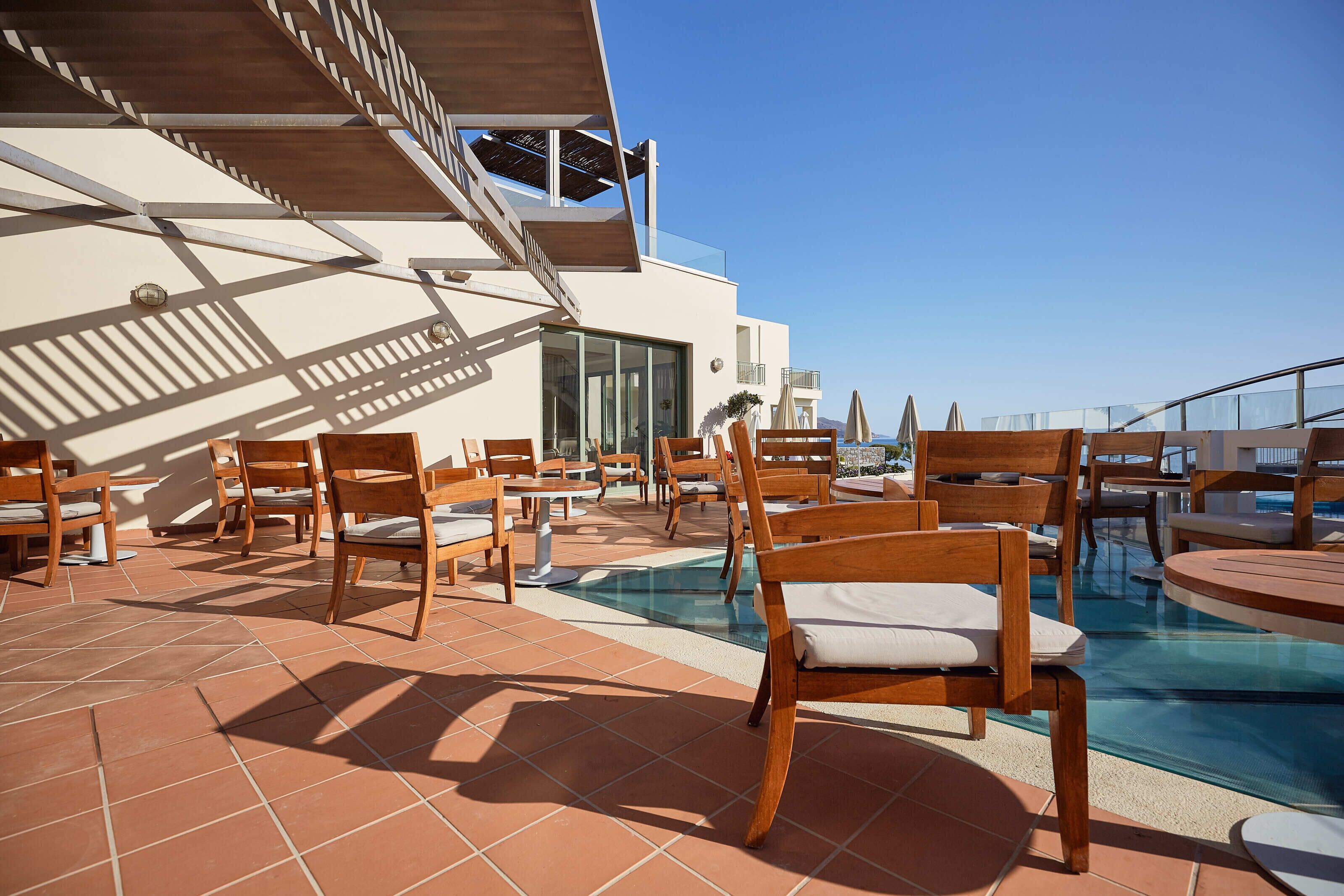 A rooftop pool area with wooden chairs and tables, a glass swimming pool, and a clear blue sky.
