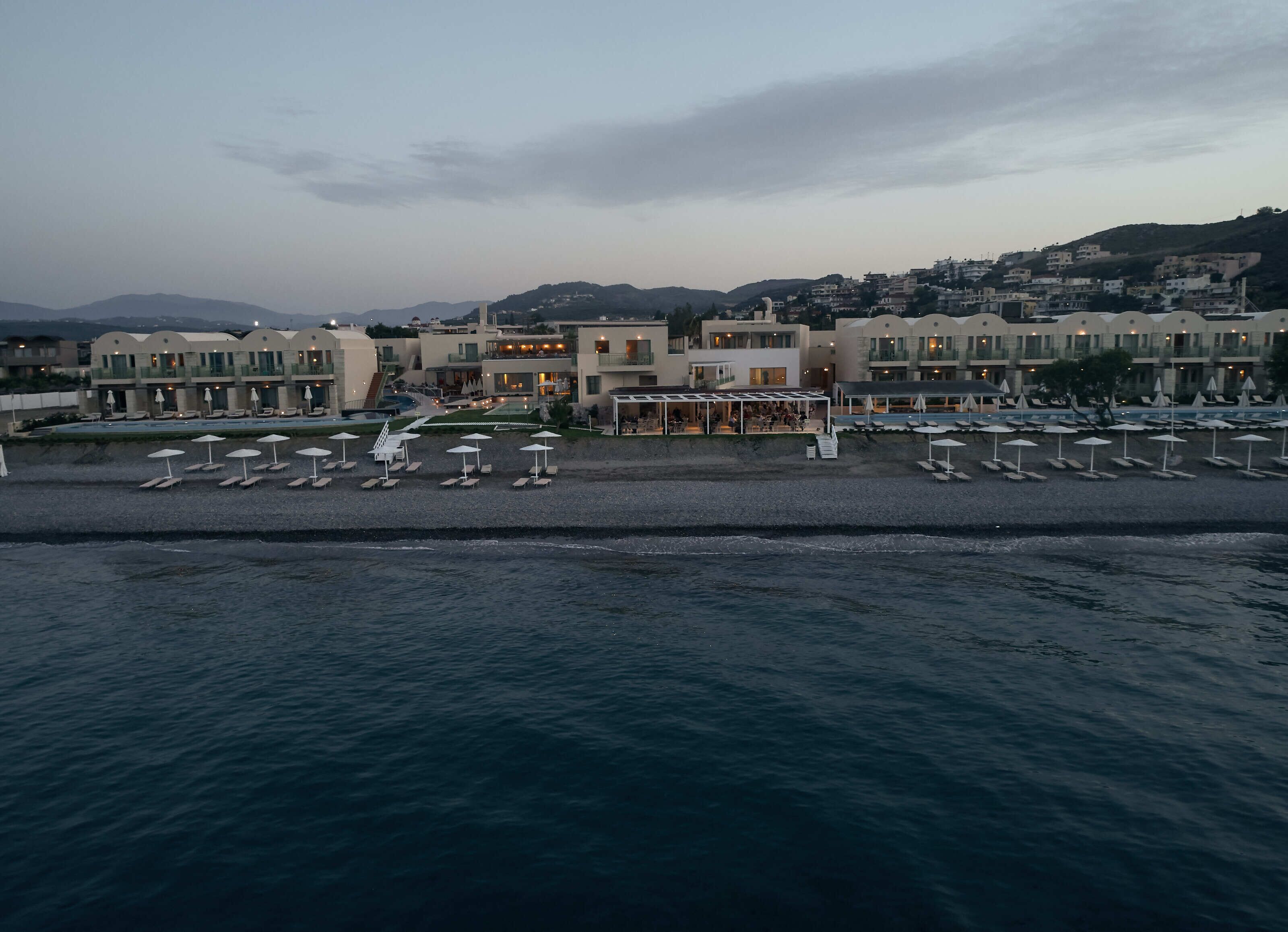 A coastal hotel with multiple buildings, a beach with umbrellas, and a view of the sea.