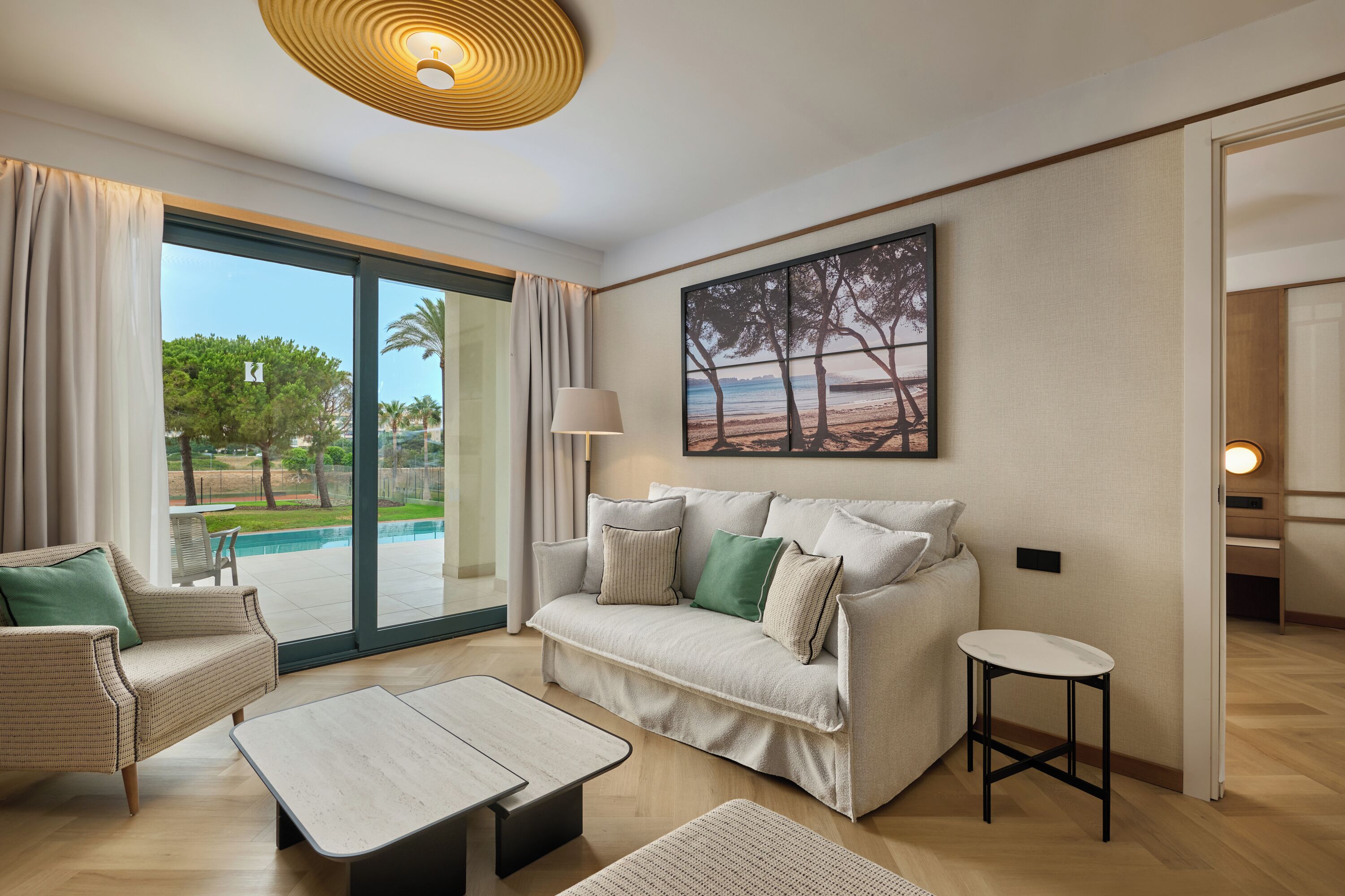 A modern living room with a sofa, a coffee table, and a side table. There is a large window with curtains, a painting on the wall, and a ceiling light fixture.