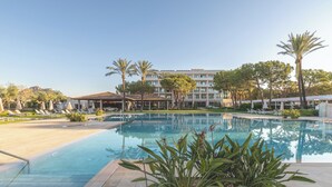 Une piscine entourée de palmiers et d’un immeuble à plusieurs étages, avec des chaises longues et des parasols à proximité.