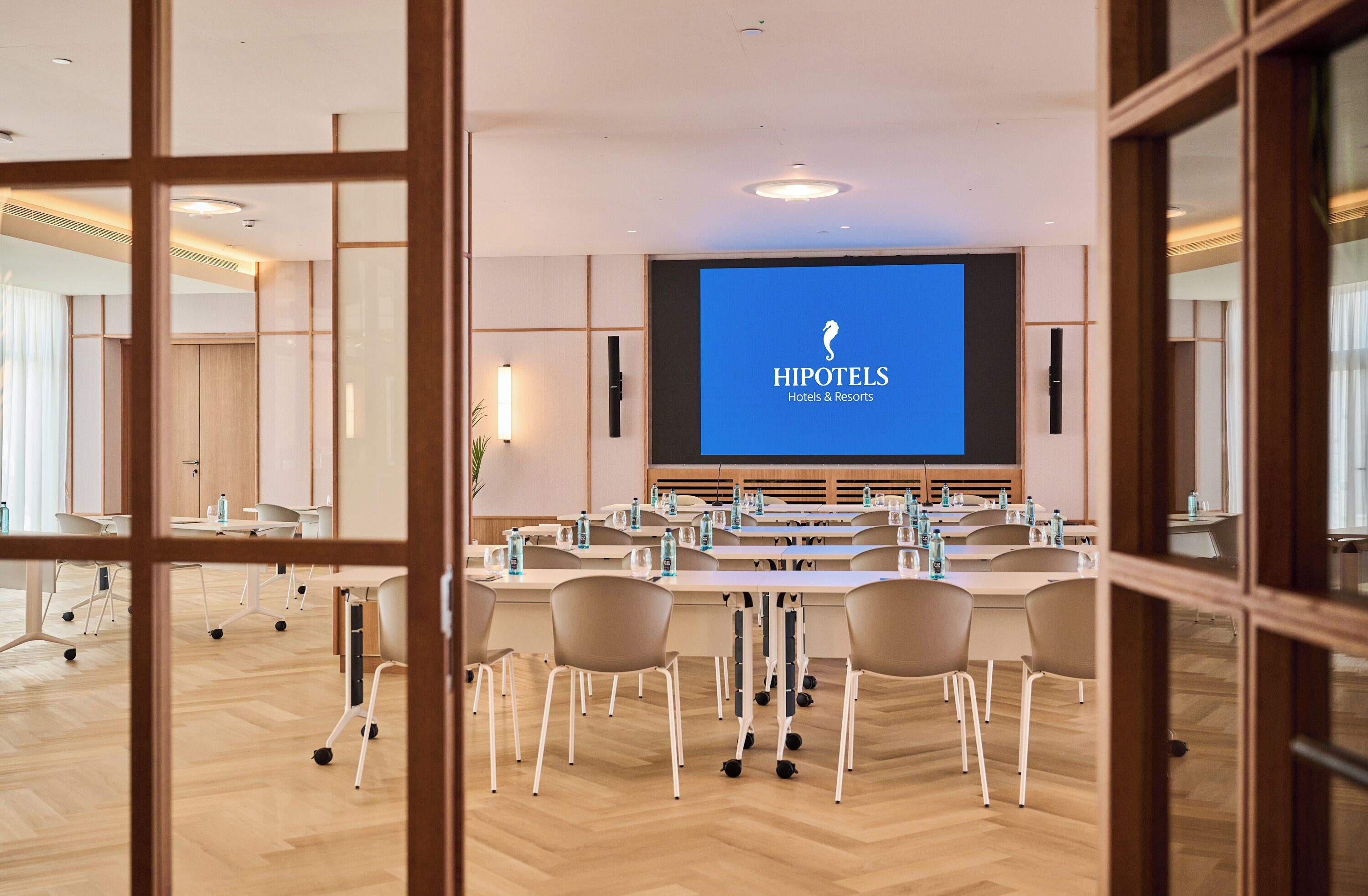 A conference room with a large screen displaying 'HIPOTELS Hotels & Resorts' logo, multiple tables with chairs, and a wooden floor.