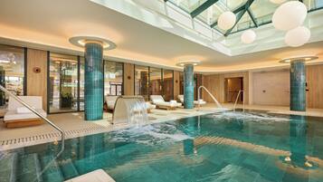 An indoor swimming pool with clear water, surrounded by lounge chairs and a glass ceiling.