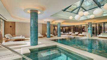 Un área de piscina interior moderna con varias zonas de estar y techo de cristal.