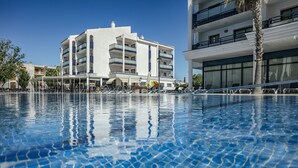 A swimming pool with clear blue water, surrounded by a white building with multiple balconies and a palm tree.