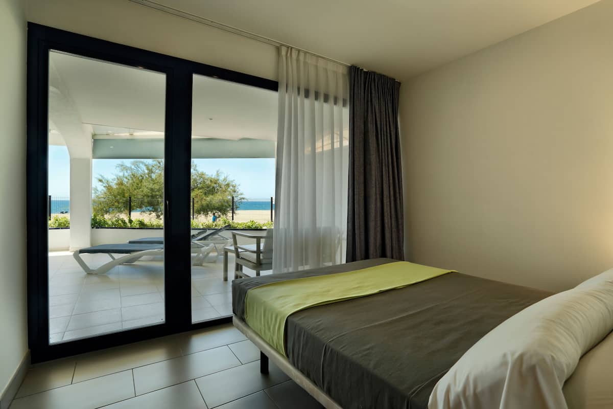 A modern bedroom with a bed, a sliding glass door leading to a balcony with a table and chairs, and a view of the outdoors.