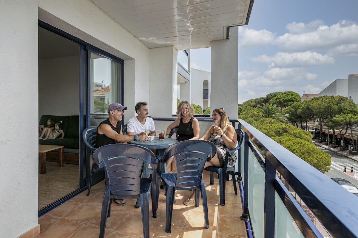 A balcony with four people seated at a table, overlooking a street with trees and buildings.